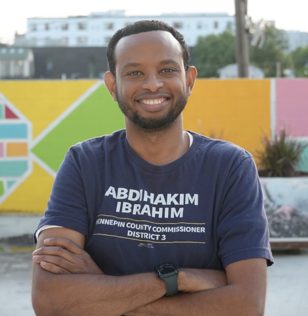 Abdihakim Ibrahim smiling in his navy campaign shirt in front of a colorful mural in District 3